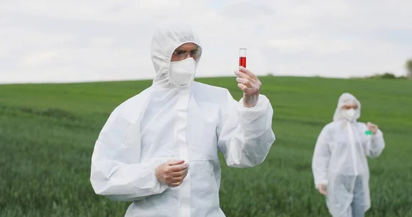 Portrait shot of Caucasian man scientist in white protective clothes and goggles holding test tube with red chemicals in green field and exploring pesticides. Woman biologist on background. — стокове фото