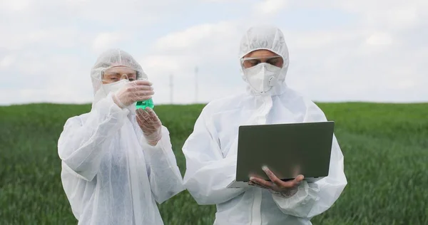 Kaukasische Wissenschaftlerin im Reagenzglas mit chemischen Pestiziden und männliche Wissenschaftlerin mit Laptop auf der grünen Wiese. Biologen-Kollegen arbeiten im Freien im Weizenfeld. — Stockfoto