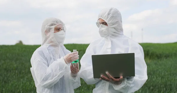 Kaukasische Wissenschaftlerin im Reagenzglas mit chemischen Pestiziden und männliche Wissenschaftlerin mit Laptop auf der grünen Wiese. Biologen-Kollegen arbeiten im Freien im Weizenfeld. — Stockfoto