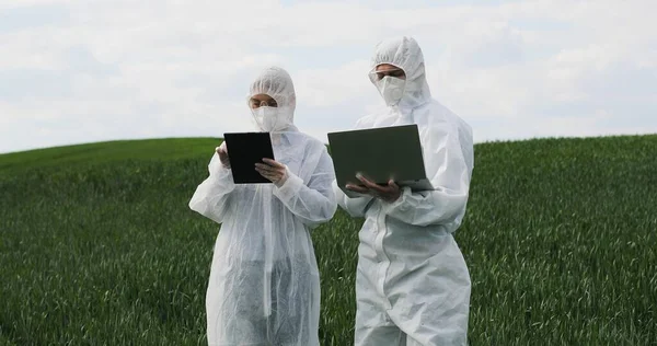 Oameni de știință caucazieni în costume de protecție care stau în câmp verde cu dispozitiv tabletă și laptop și vorbesc. Bărbat și femeie biolog care lucrează în marjă cu recolta . — Fotografie, imagine de stoc