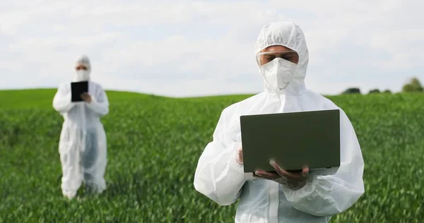 Homem biólogo agricultor caucasiano em traje protetor branco e óculos de pé no campo verde e trabalhando no computador portátil. Homem ecologista agrícola e cientista explorando colheita . — Fotografia de Stock