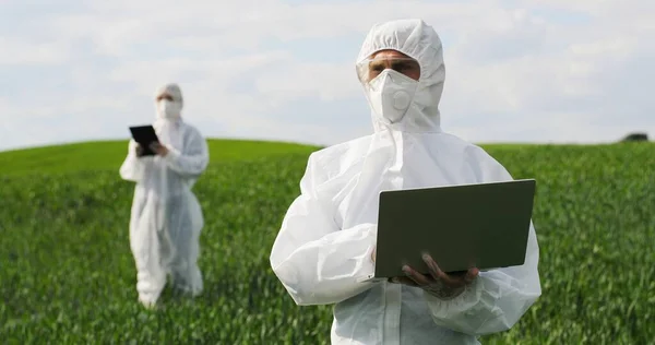 Männlicher kaukasischer Bauernbiologe in weißem Schutzanzug und Schutzbrille, der auf der grünen Wiese steht und am Laptop arbeitet. Mann Agrarökologe und Wissenschaftler erforscht Ernte. — Stockfoto