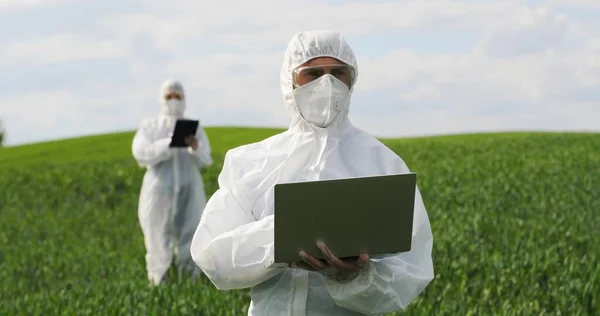 Männlicher kaukasischer Bauernbiologe in weißem Schutzanzug und Schutzbrille, der auf der grünen Wiese steht und am Laptop arbeitet. Mann Agrarökologe und Wissenschaftler erforscht Ernte. — Stockfoto