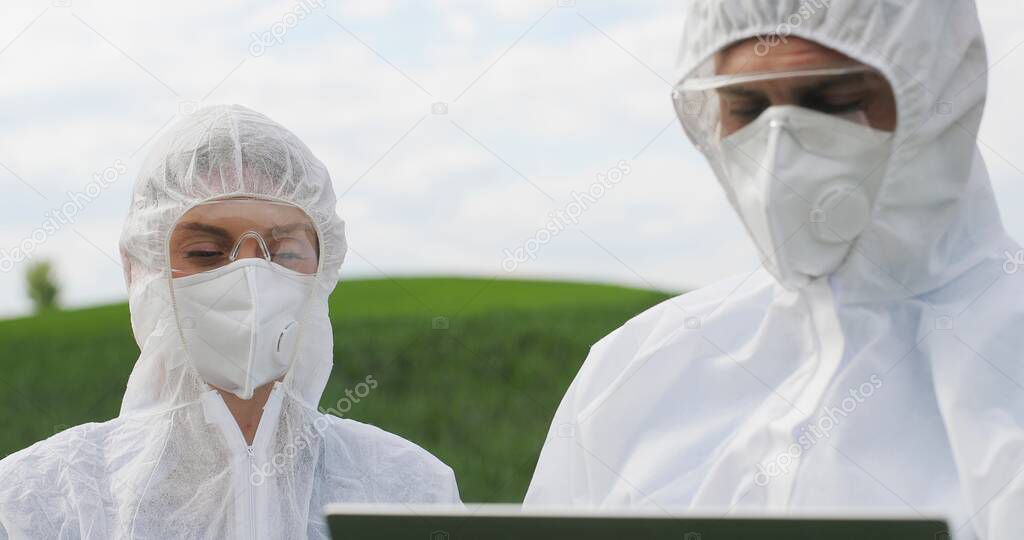 Close up of Caucasian female and male farmers researchers holding test tube with chemicals pesticides and laptop computer in green field. Biologists co-workers working and talking in wheat margin.