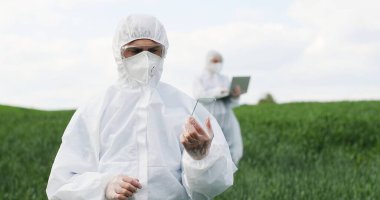 Glass futuistic smartphone in hands of male ecologist standing in green field in summer. Hi-tech technology of transparent device. Man in protective suit tapping on phone of future. Augmented reality.