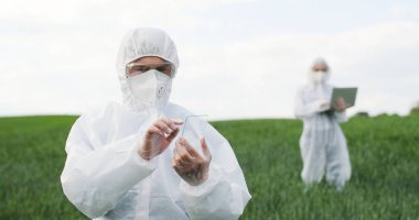 Glass futuistic smartphone in hands of male ecologist standing in green field in summer. Hi-tech technology of transparent device. Man in protective suit tapping on phone of future. Augmented reality.