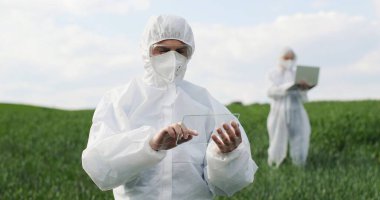 Caucasian man scientist in protective costume standing in field, tapping on glass as on screen of futuristic device. Screentouch concept. Future technology. Male fermer using transparent computer.