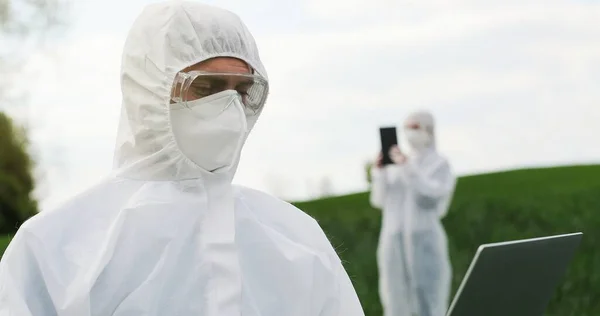 Біолог чоловічої кавказької ферми в білому захисному костюмі, масці і окулярах, що стоять на зеленому полі і працюють на ноутбуці. Вивчення урожаю і шкідників людиною - екологом. — стокове фото