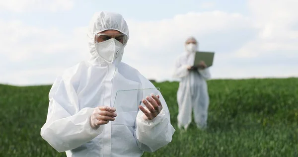 Caucasian man scientist in protective costume standing in field, tapping on glass as on screen of futuristic device. Screentouch concept. Future technology. Male fermer using transparent computer. — Stockfoto