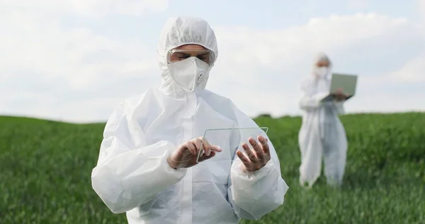 Caucasian man scientist in protective costume standing in field, tapping on glass as on screen of futuristic device. Screentouch concept. Future technology. Male fermer using transparent computer. — Stockfoto