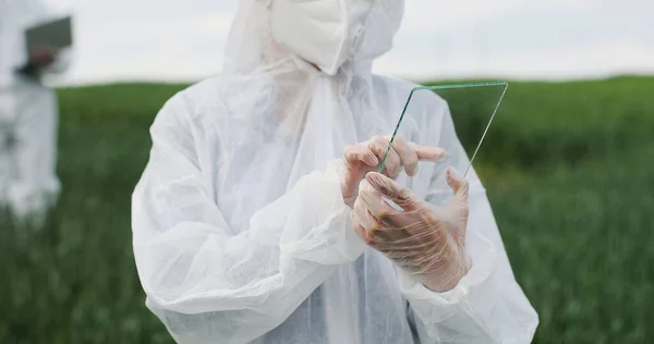 Närbild av kvinnliga bonde ekolog i skyddsdräkt knacka på glas transparent skärm. Futuristisk apparat i fält. Pekskärm med högteknologi. Kvinnlig forskare med framtidens surfplatta i händerna. — Stockfoto