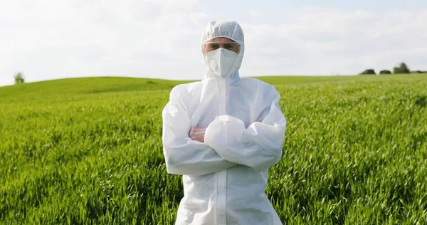 Portrait of Caucasian male farmer ecologist in white protective costume, mask and goggles standing in green field and turning face to camera. Man scientist and biologist posing in margin with harvest. — Stockfoto