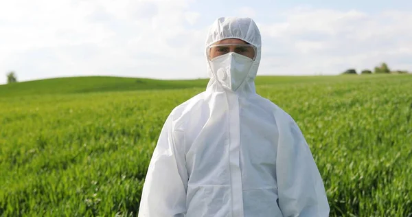 Portrait of Caucasian male farmer ecologist in white protective costume, mask and goggles standing in green field and turning face to camera. Man scientist and biologist posing in margin with harvest. — стокове фото