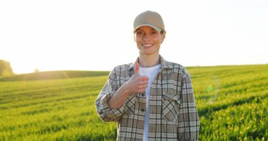 Portrait shot of beautiful young Caucasian woman in hat standing in green field and smiling cheerfully to camera. Female farmer looking straight with smile outdoor in summer.