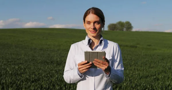 Caucasian beautiful female ecologist scientist in white gown walking in green field and holding tablet device. Woman researcher, biologist strolling in margin of harvest while using tablet computer. — Stock Photo, Image