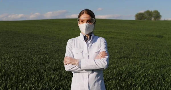 Portrait of Caucasian beautiful female ecologist scientist in white gown, mask and googles standing in green field and looking at camera with crossed hands. Woman researcher, biologist in margin. — Stockfoto
