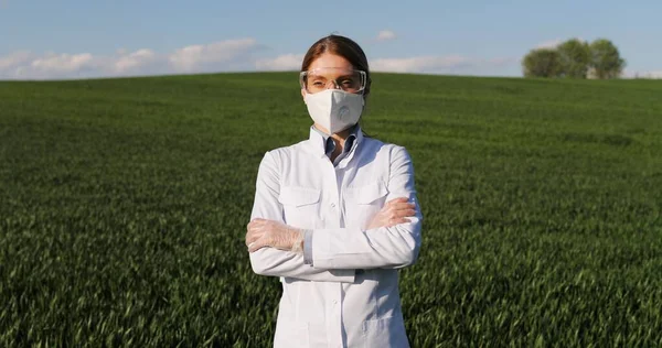 Portrait of Caucasian beautiful female ecologist scientist in white gown, mask and googles standing in green field and looking at camera with crossed hands. Woman researcher, biologist in margin. — Stockfoto