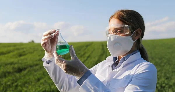 Close up of Caucasian beautiful female ecologist scientist in white gown, mask and googles standing in green field and looking at chemicals in test tube. Woman researcher in margin studying pesticides — Stockfoto