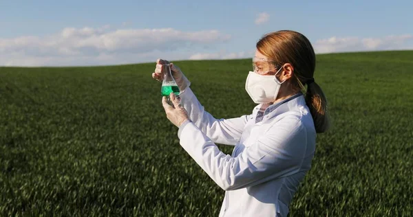 Caucasiana cientista ecologista bonita do sexo feminino em vestido branco, máscara e óculos em pé no campo verde e olhando para produtos químicos em tubo de ensaio. Mulher pesquisadora e bióloga em margem estudando agrotóxicos . — Fotografia de Stock