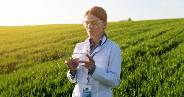 Caucasian beautiful female ecologist scientist in white gown, mask and goggles standing in green field and working on tablet device. Woman researcher, biologist in margin using tablet computer. — Stock Photo, Image