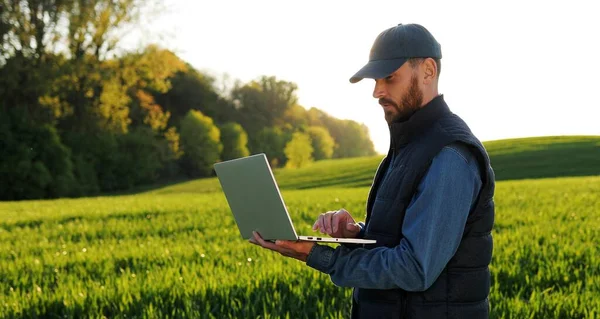 Giovane agricoltore maschio caucasico in cappello in piedi in campo e digitando sulla tastiera del computer portatile. Uomo attraente utilizzando dispositivo a margine in estate e la navigazione online. Concetto agricolo . — Foto Stock