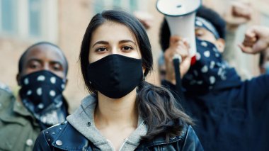 Close up of young Caucasian beautiful woman in mask looking straight at camera outdoor at street riot. Portrait of girl with male Afrcan Americans on background. Female protester at manifestation. clipart