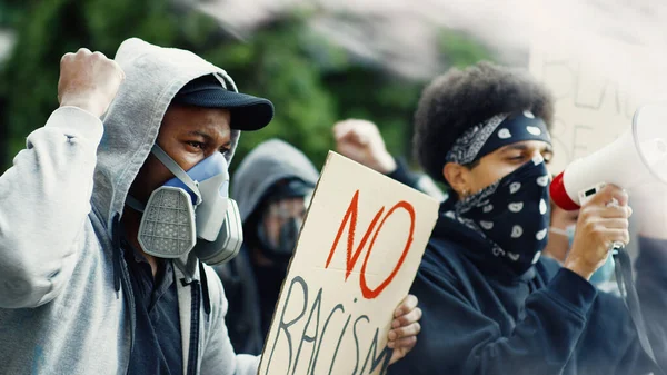 Mixed-races males protesters in respirators and masks protesting with colorful smoke and posters against racism and police brutality in USA. Multiethnic men fighting for equal rights at street riot.