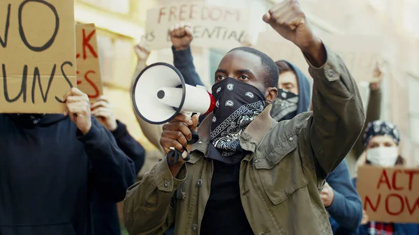 Afrikai-amerikai jóképű férfi sállal az arcán, mint egy maszk, aki hangosbeszélőben sikoltozik az emberi jogok elleni tiltakozásért a szabadban. Emberek egy csoportja tiltakozik az utcán. Erőszak elleni küzdelem. — Stock Fotó
