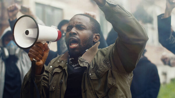 African-American young handsome man screaming in megaphone at protest for human rights outdoors in smoke. Group of people protesting at street. Strike against violence.