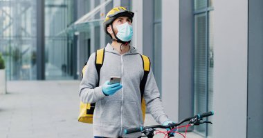 Tıbbi maskeli teslimatçı, eldivenler ve casque caddede bisikletini sürüyor, cep telefonunu dinliyor, kuryeye telefon açıyor..