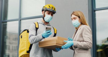 Tıbbi maskeli genç bir erkek kurye karton kutuları sokaktaki bir kadına getiriyor ve paketleri kontrol etmek için telefonu dinliyor. Yemek servisi konsepti