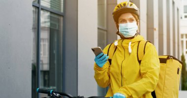 Bisiklet sürmek için bisiklet ve yiyecek teslimatı yapan sarı termal çantalı ve kasklı kadın kurye portresi. Teslimatçı akıllı telefon kullanıyor.
