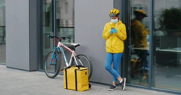 Female delivery worker in medical mask standing at bicycle outdoor and tapping or scrolling on smartphone.