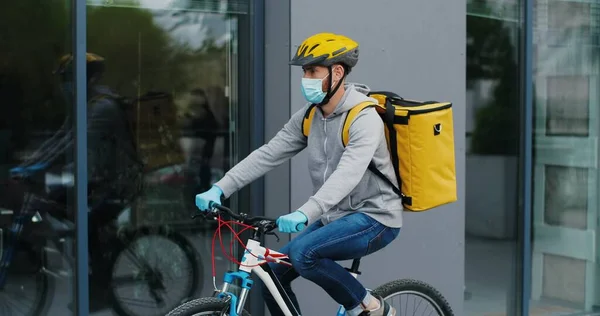 Young courier in medical mask delivering food with yellow thermal backpack, riding a bicycle in the city. Food delivery service concept