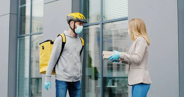 Portrait of delivery man with yellow thermal bag for food delivery in medical mask holding pizza boxes in front house and woman accepting a delivery of pizza boxes from courier.