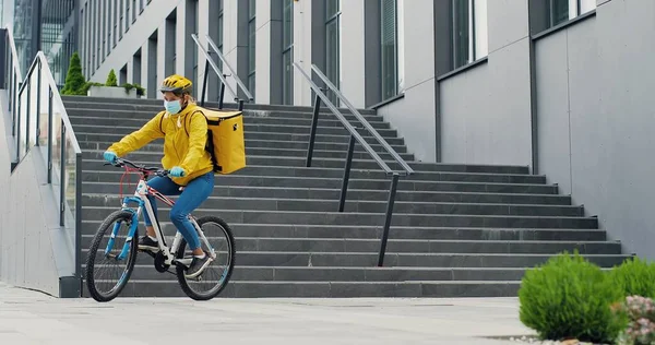 Female courier in medical mask delivering food with yellow thermal backpack, riding a bicycle in the city. Food delivery service concept
