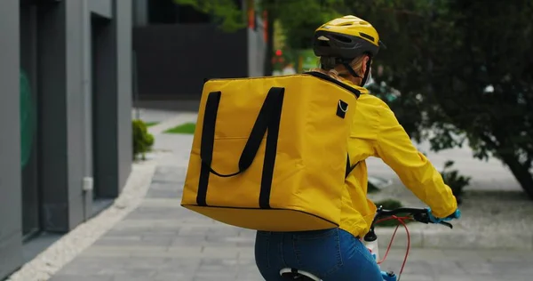 Female courier in medical mask delivering food with yellow thermal backpack, riding a bicycle in the city. Food delivery service concept