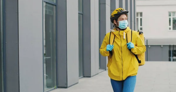 Portrait of beautiful woman delivery worker in medical mask and helmet for riding bike walking the street. Pretty female courier with backpack. Food delivery concept