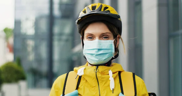 Portrait of beautiful woman delivery worker in medical mask and helmet for riding bike standing at street. Pretty female courier with backpack. Food delivery concept