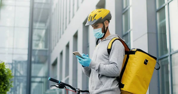 Portrait of male courier in medical mask and helmet for riding bike on bicycle delivering food. Deliveryman using smartphone