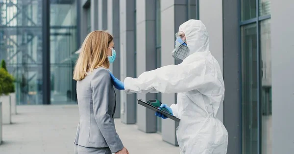 Lavoratrice in maschera che va al lavoro e uomo in completo bianco e respiratore che misura la sua temperatura, lasciandola andare. Permesso di passaggio al business center per i visitatori. Pandemia di Covid-19. Permesso di entrare. — Foto Stock