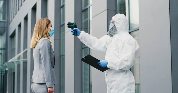 Lavoratrice in maschera che va al lavoro e uomo in completo bianco e respiratore che misura la sua temperatura, lasciandola andare. Permesso di passaggio al business center per i visitatori. Pandemia di Covid-19. Permesso di entrare. — Foto Stock