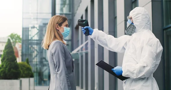 Female office worker in mask going to work and man in white suit and respirator measuring her temperature, letting go. Allowing pass to business center for visitor. Covid-19 pandemic. Permit to enter.