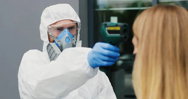 Lavoratrice in maschera che va al lavoro e uomo in completo bianco e respiratore che misura la sua temperatura, lasciandola andare. Permesso di passaggio al business center per i visitatori. Pandemia di Covid-19. Permesso di entrare. — Foto Stock