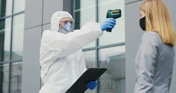 Lavoratrice in maschera che va al lavoro e uomo in completo bianco e respiratore che misura la sua temperatura, lasciandola andare. Permesso di passaggio al business center per i visitatori. Pandemia di Covid-19. Permesso di entrare. — Foto Stock