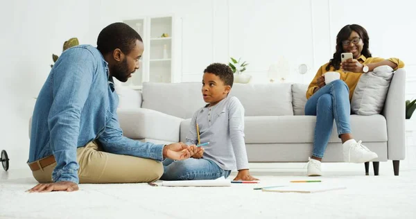 Gelukkig Afrikaans-Amerikaans gezin dat samen tijd doorbrengt thuis. Vrolijke Afrikaanse vader spelen met kleine zoon, jonge moeder met behulp van smartphone. — Stockfoto