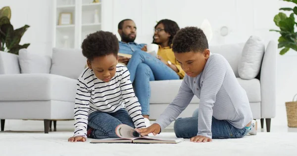 Gelukkig Afrikaans-Amerikaans gezin dat samen tijd doorbrengt thuis. Ouders ontspannen samen op de bank in de woonkamer, kleine kinderen broer en zus lezen boek thuis, familie tijd concept — Stockfoto