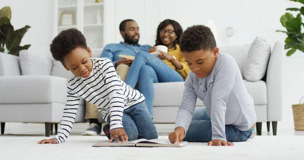 Gelukkig Afrikaans-Amerikaans gezin dat samen tijd doorbrengt thuis. Ouders ontspannen samen op de bank in de woonkamer, kleine kinderen broer en zus lezen boek thuis, familie tijd concept — Stockfoto