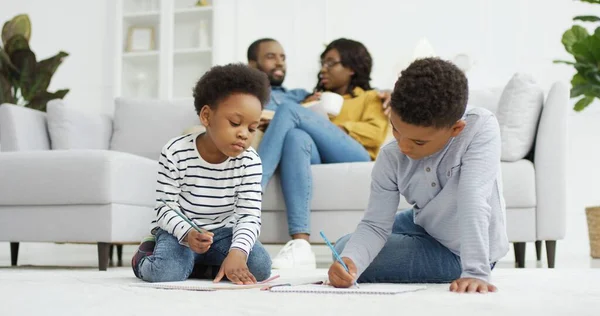 Gelukkig Afrikaans-Amerikaans gezin dat samen tijd doorbrengt thuis. Ouders ontspannen samen op de bank in de woonkamer, kleine kinderen broer en zus tekenen thuis. — Stockfoto