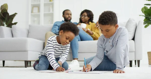 Gelukkig Afrikaans-Amerikaans gezin dat samen tijd doorbrengt thuis. Ouders ontspannen samen op de bank in de woonkamer, kleine kinderen broer en zus tekenen thuis. — Stockfoto